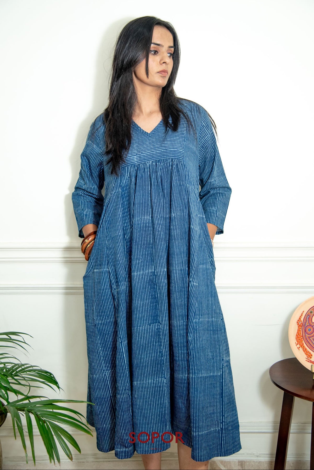 A woman modeling a loose-fitting, long-sleeved blue dress with a textured vertical stripe pattern, standing against a white wall.